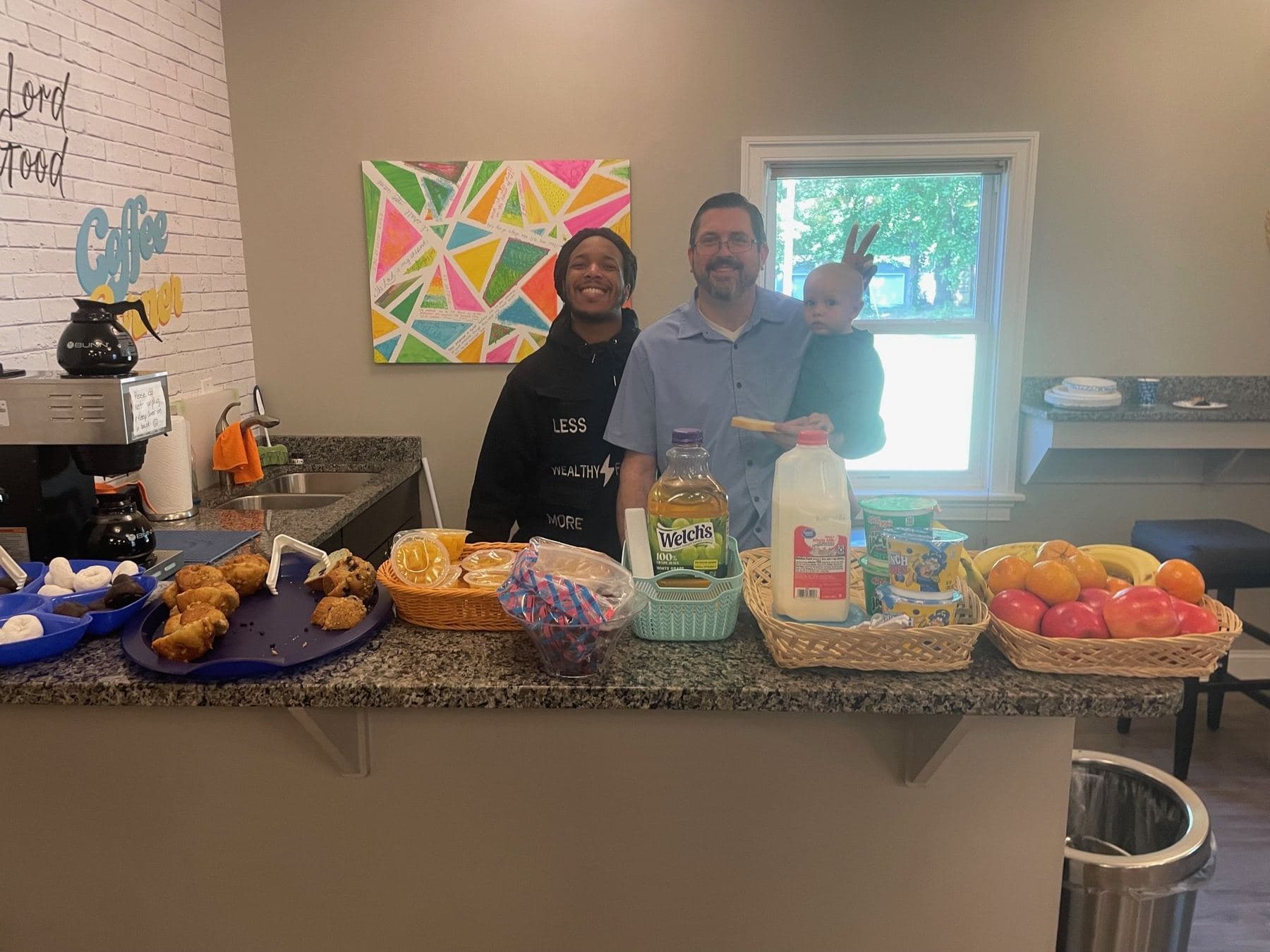 Sunday breakfast laid out along the Coffee Corner counter — fruit, pastries, and cereal — with three members behind it, one holding a baby