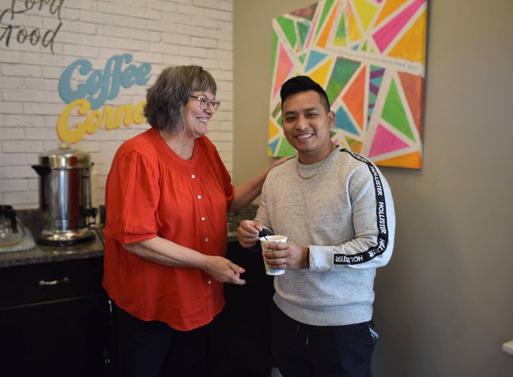 Two church members laughing together over coffee beneath the hand-painted Coffee Corner sign