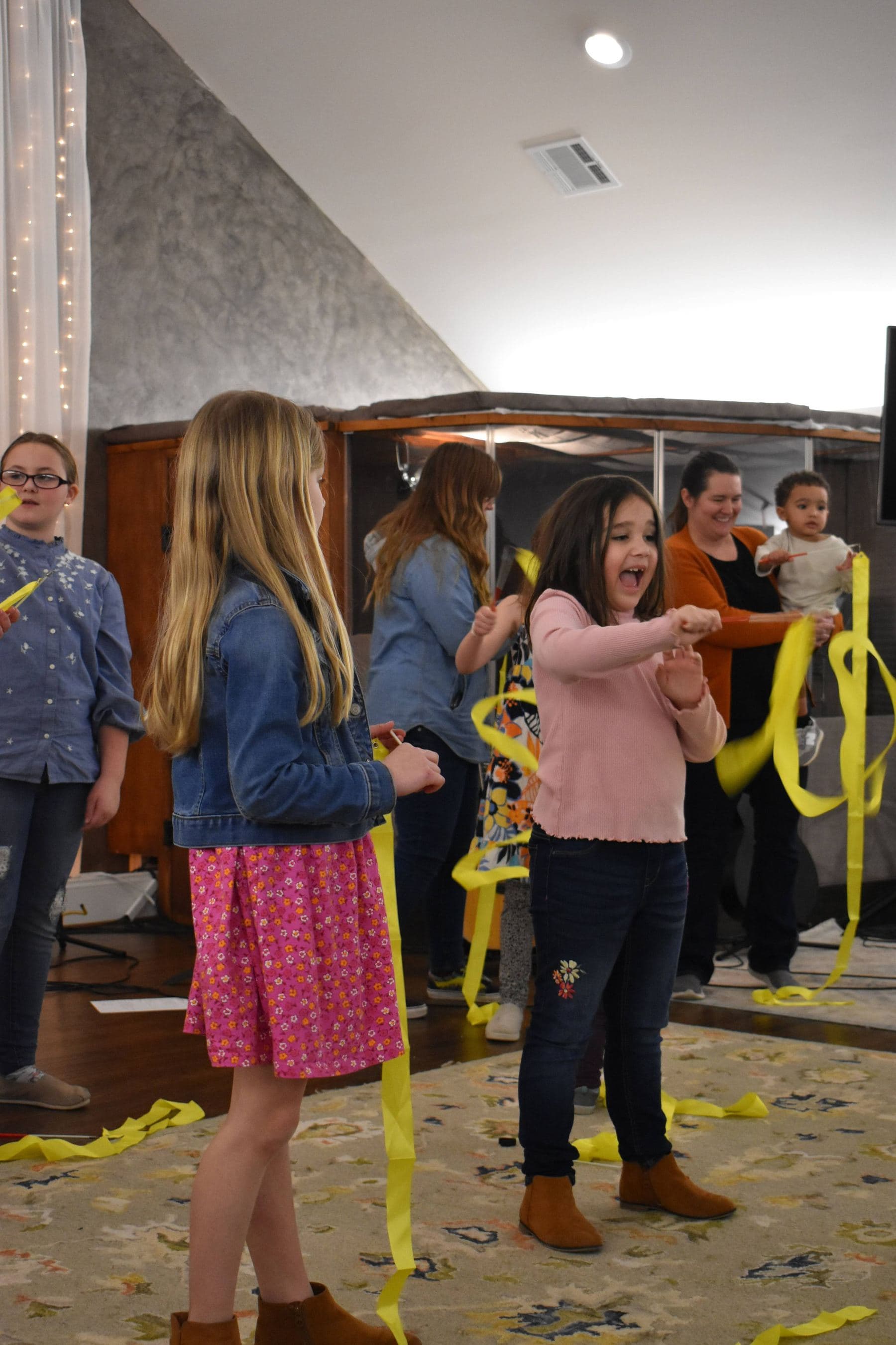 Children playing with long yellow streamers in the fellowship hall, one girl mid-laugh