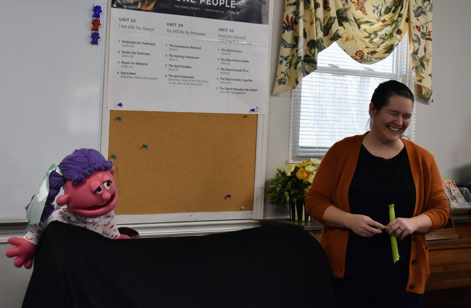 A children's ministry teacher laughing mid-lesson with a puppet on the table beside her