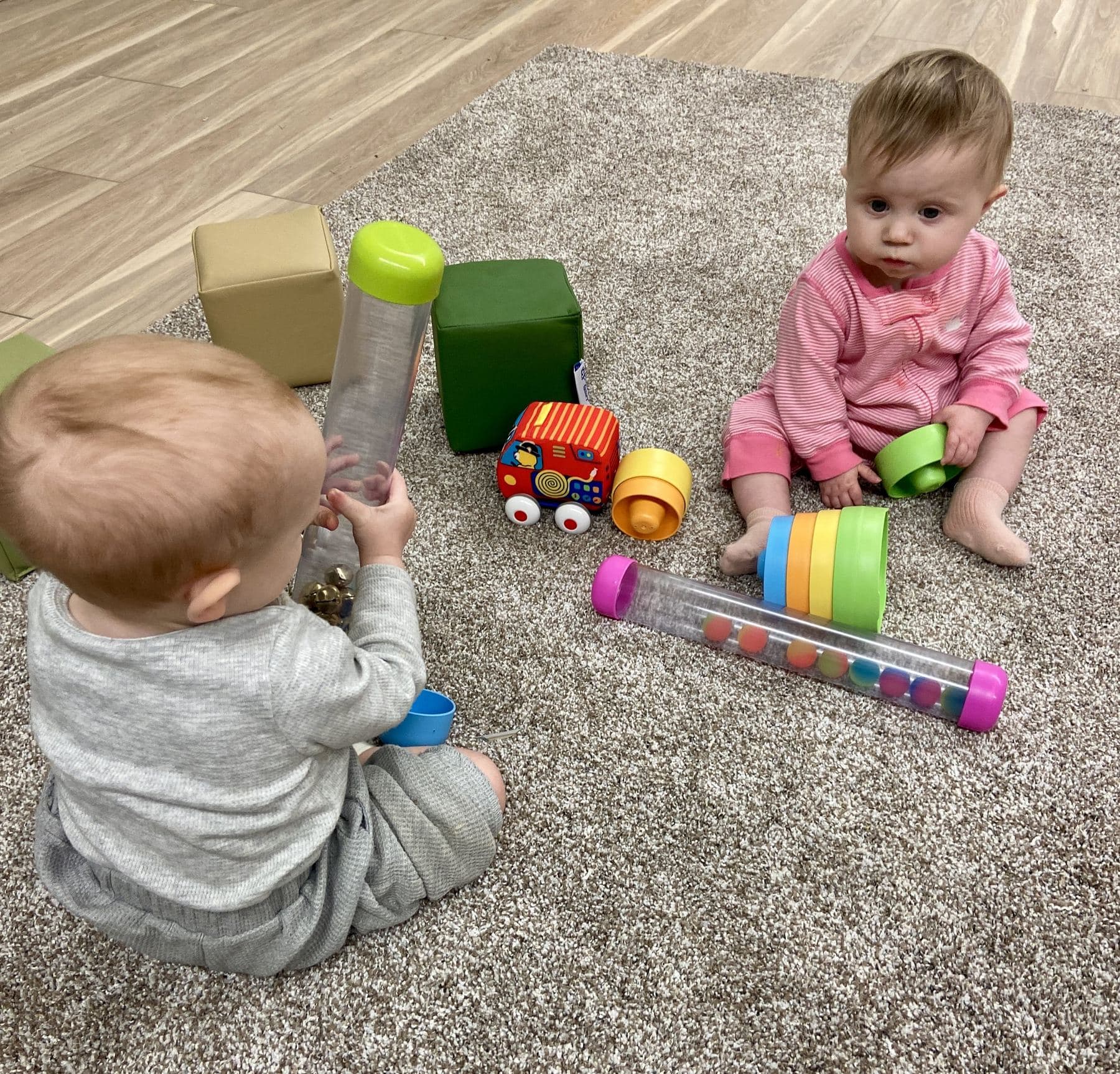 Two babies on the nursery carpet, one shaking a sensory tube and one stacking rings