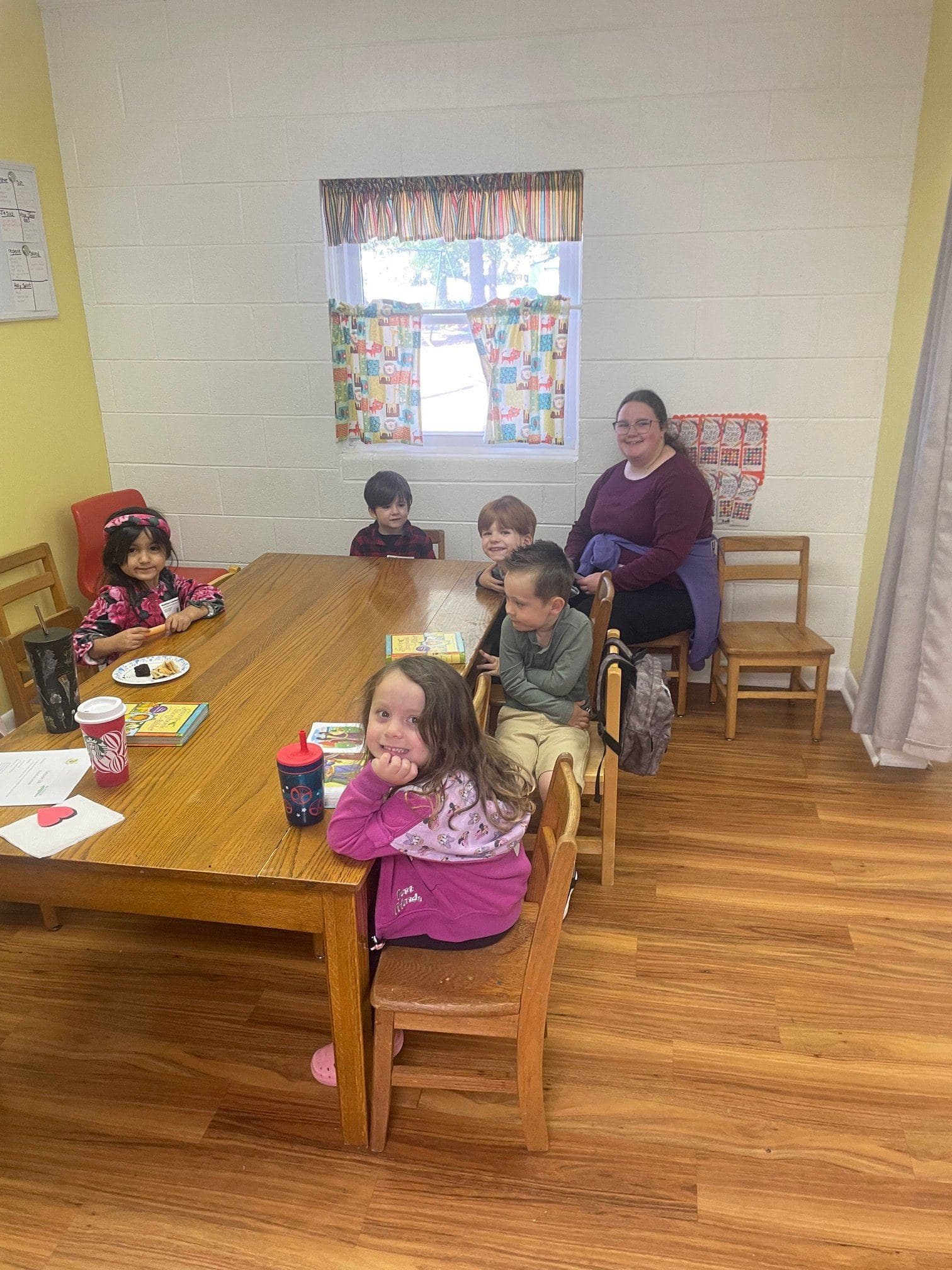Preschoolers at the snack table in their classroom with their teacher, one little girl grinning at the camera