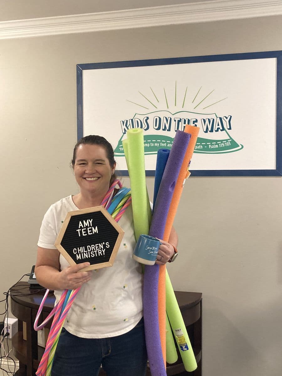 Amy Teem in the children's wing, holding a letterboard reading Amy Teem, Children's Ministry, with an armful of pool noodles