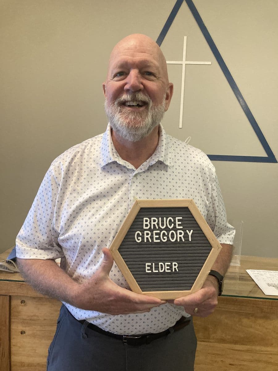 Bruce Gregory holding a letterboard reading Bruce Gregory, Elder, in front of the church's painted cross-and-triangle wall mark