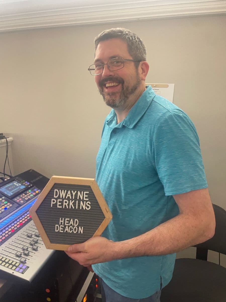 Dwayne Perkins at the media desk holding a letterboard reading Dwayne Perkins, Head Deacon