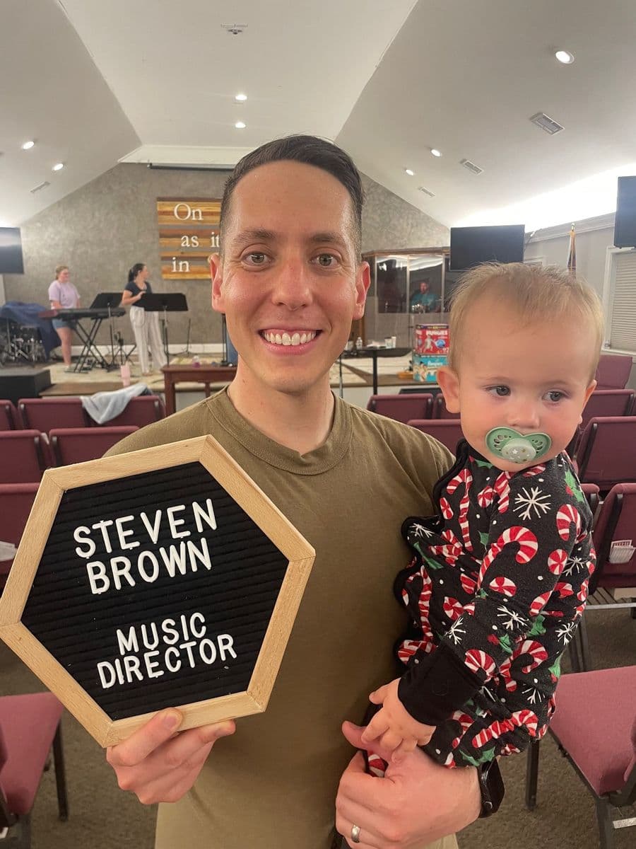 Steven Brown in the sanctuary holding his baby and a letterboard reading Steven Brown, Music Director