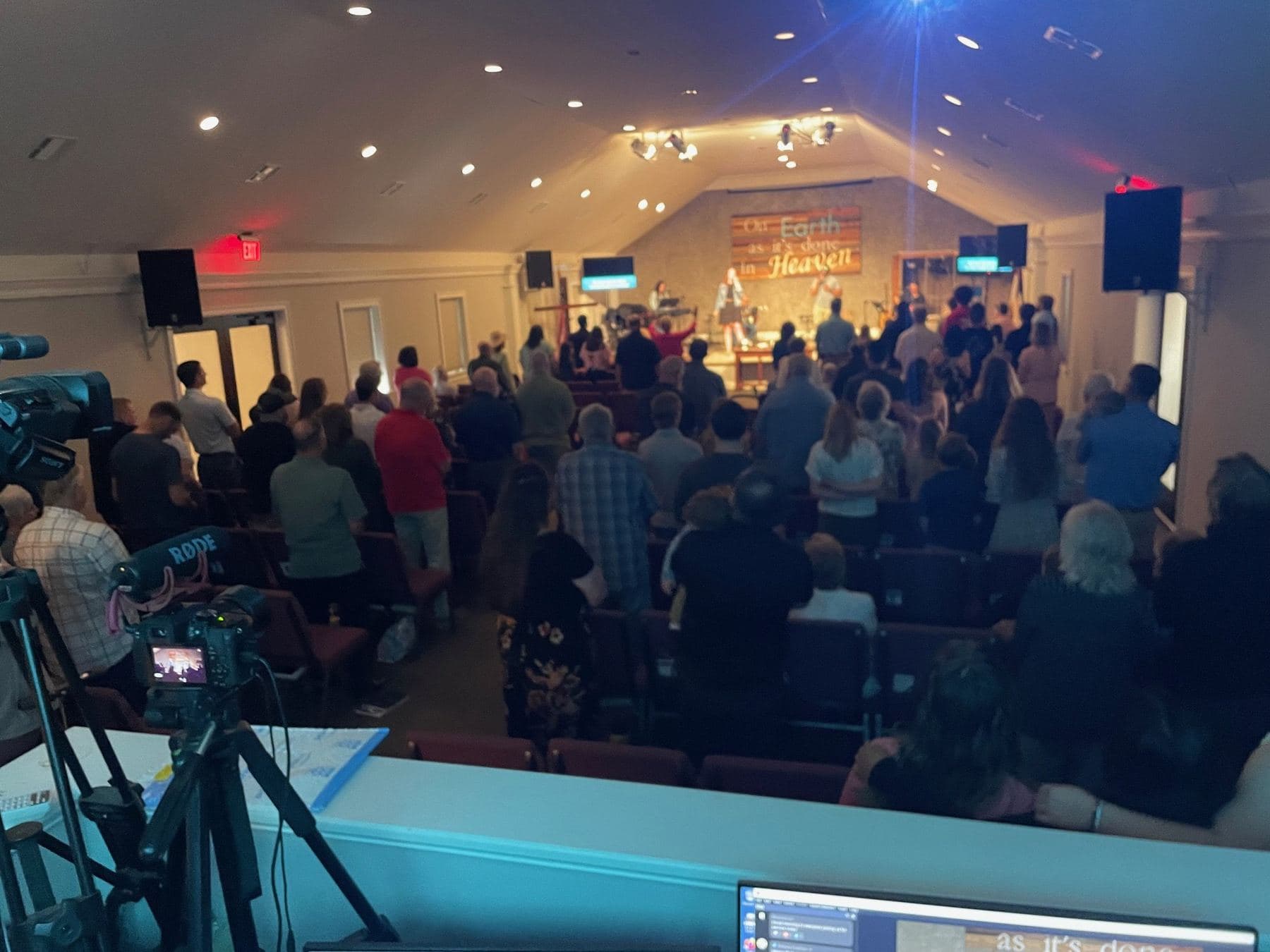 A full Sunday morning service from the back of the room, the congregation standing to sing under the sanctuary's peaked ceiling