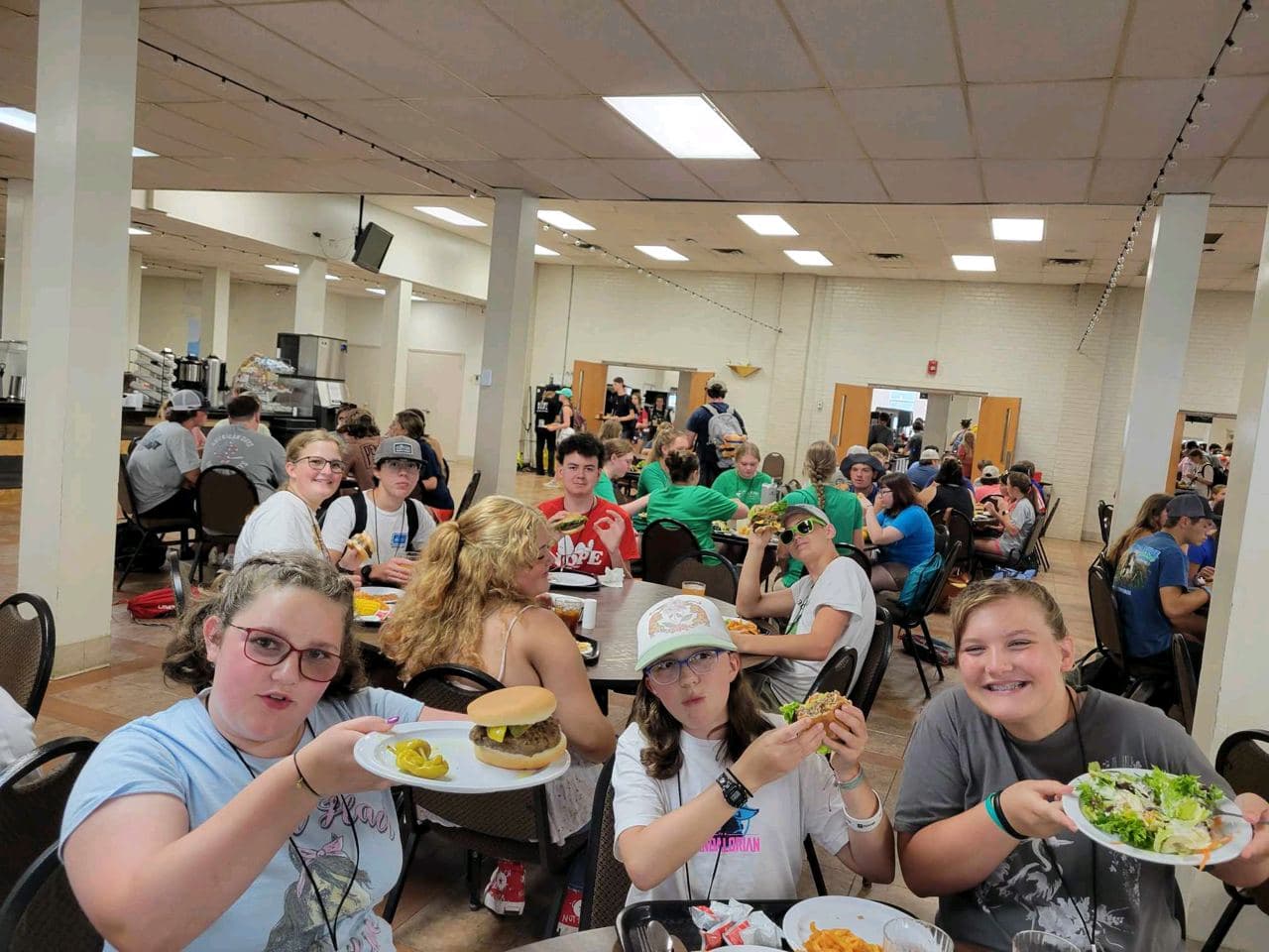 Students at long tables in the camp dining hall holding up their lunch plates