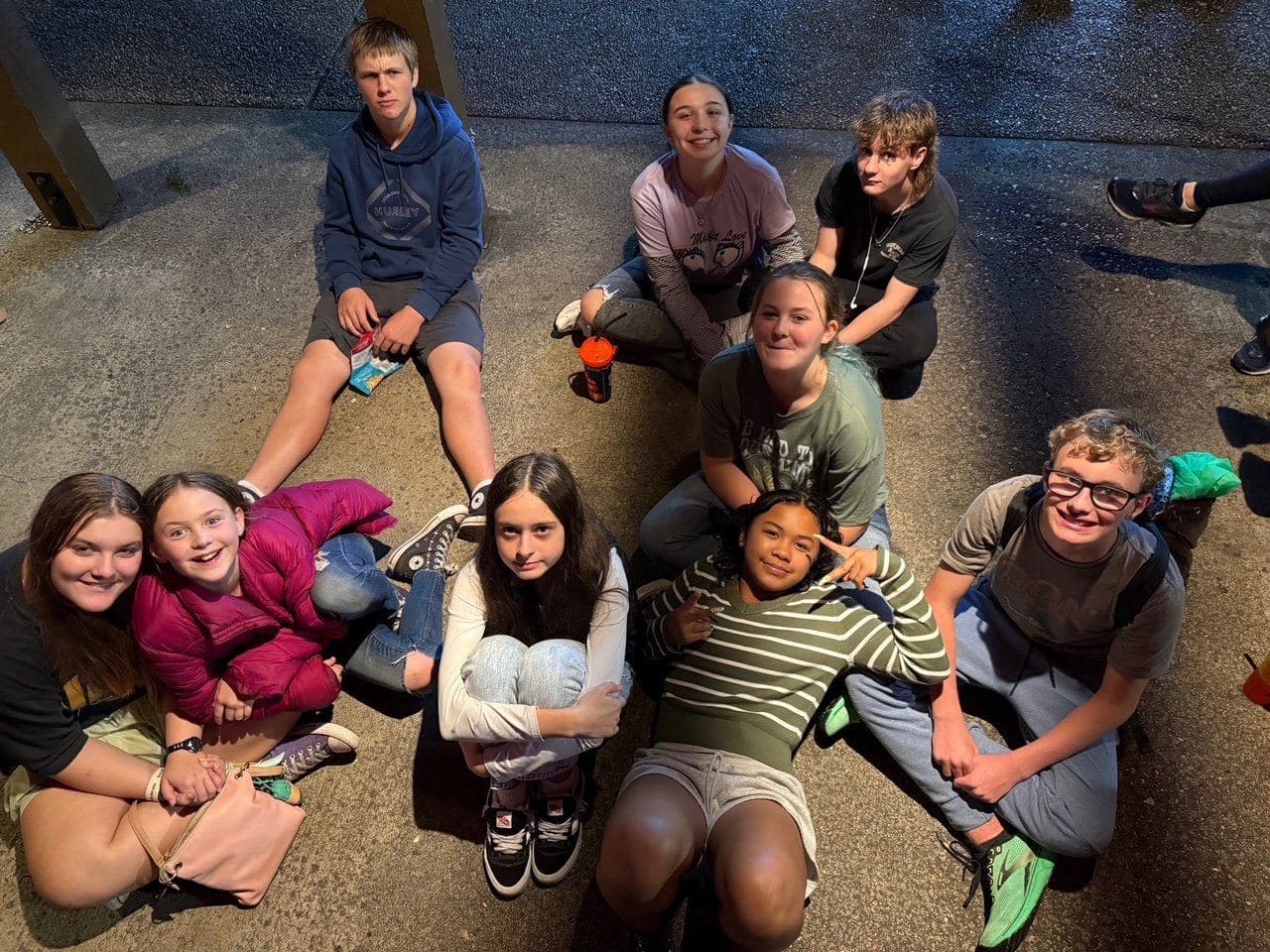 Nine teenagers sitting together on the pavement outside on a summer evening, looking up at the camera